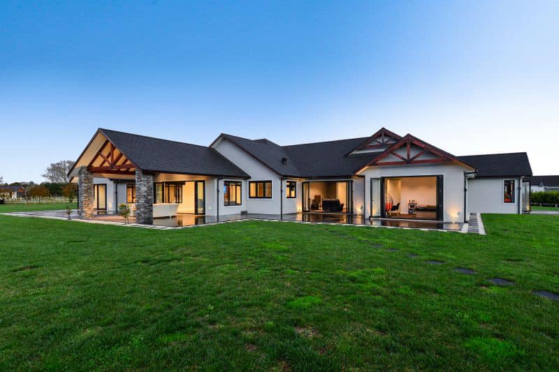 Modern single-level home with large windows, open gables, and a spacious lawn, photographed at dusk with warm interior lighting.