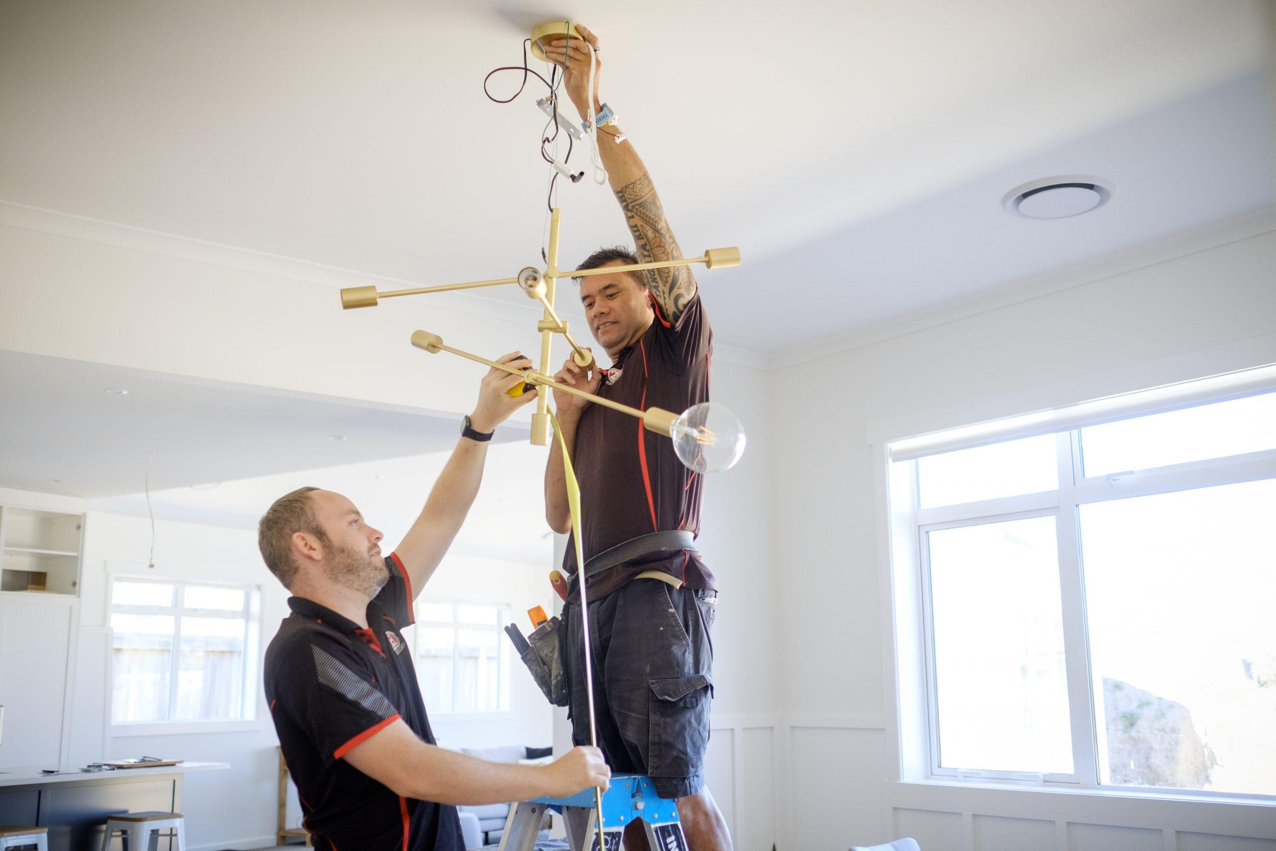 Two electricians from 2E Electrical installing a modern gold pendant light fixture in a bright residential living space.
