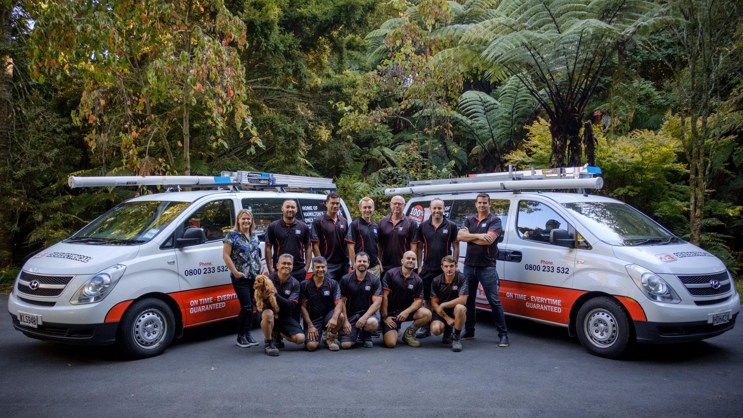 2E Electrical team posing outdoors with company vans, showcasing their full crew and branded fleet.