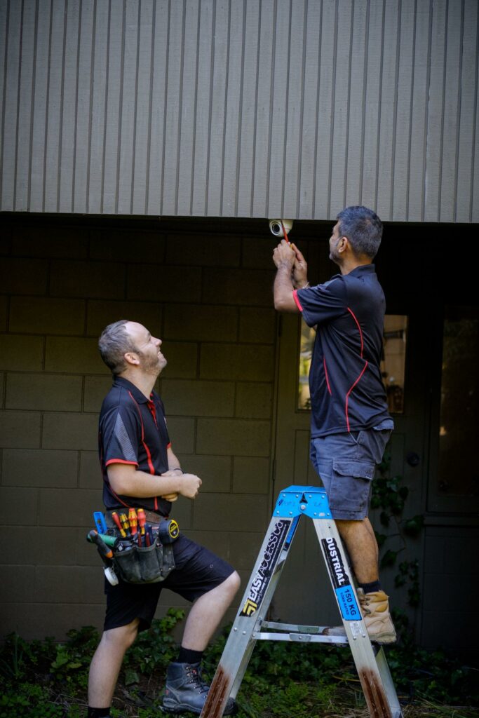 Two electricians in 2E Electrical uniforms working on an outdoor lighting fixture, one on a ladder while the other stands beside him with tools.